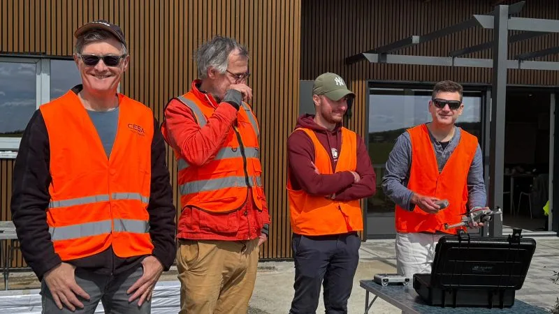 Groupe de stagiaires en gilets haute visibilité sur le terrain, drone et radiocommande en main, mallette de transport ouverte sur la table — formation de télépilote de drone Dronexperts